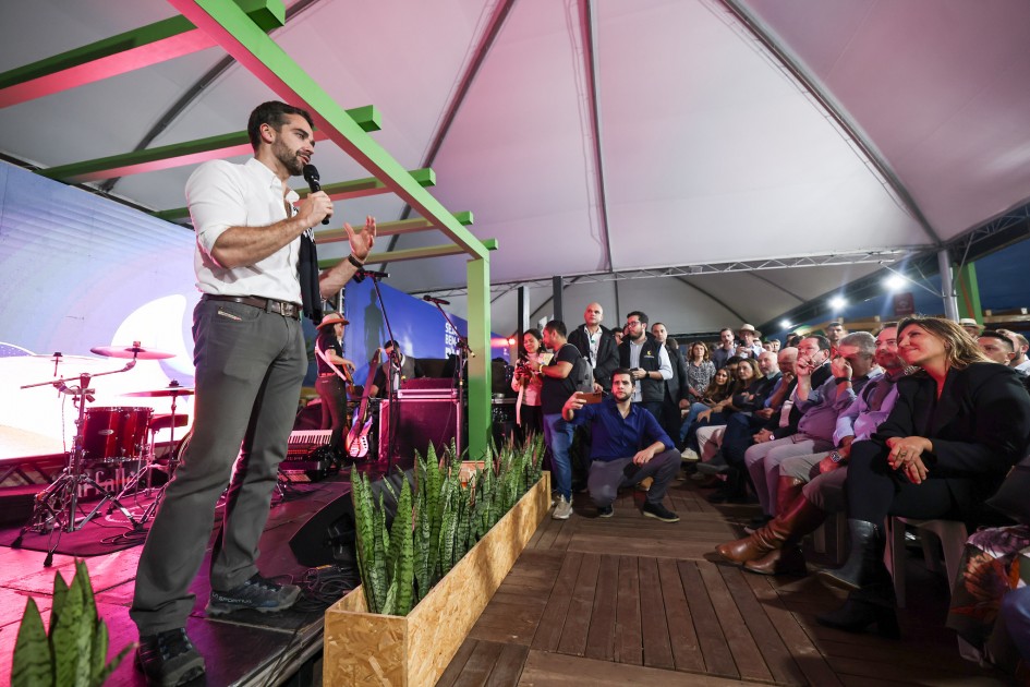 Em um palco coberto por tenda branca, um homem de camisa branca e calça cinza fala ao microfone, de mangas dobradas, com um tecido preto de letras brancas apoiado sobre um ombro. Ele gesticula diante de uma plateia numerosa e atenta, com pessoas sentadas e algumas em pé registrando a cena; um homem agachado fotografa no corredor. À esquerda, aparecem bateria, amplificadores e músicos; atrás do orador há um telão iluminado. No primeiro plano, um canteiro de espadas-de-São-Jorge e uma estrutura com vigas verdes contornam o palco.