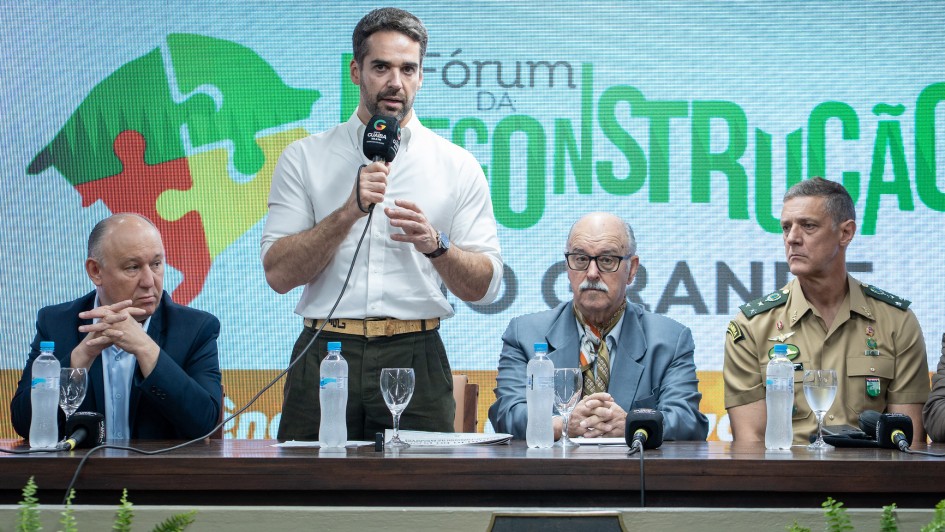 Em um evento oficial, um homem de camisa branca e mangas dobradas fala ao microfone em pé atrás de uma mesa de autoridades. Ao lado dele, três homens estão sentados, entre eles um de terno claro com lenço no pescoço e outro com farda militar bege com insígnias. Sobre a mesa há microfones, taças e garrafas de água. Ao fundo, um telão exibe a frase “Fórum da Reconstrução” com ilustração de peças de quebra-cabeça; na parte inferior da imagem, há samambaias decorativas.