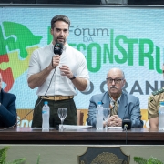 Em um evento oficial, um homem de camisa branca e mangas dobradas fala ao microfone em pé atrás de uma mesa de autoridades. Ao lado dele, três homens estão sentados, entre eles um de terno claro com lenço no pescoço e outro com farda militar bege com insígnias. Sobre a mesa há microfones, taças e garrafas de água. Ao fundo, um telão exibe a frase “Fórum da Reconstrução” com ilustração de peças de quebra-cabeça; na parte inferior da imagem, há samambaias decorativas.