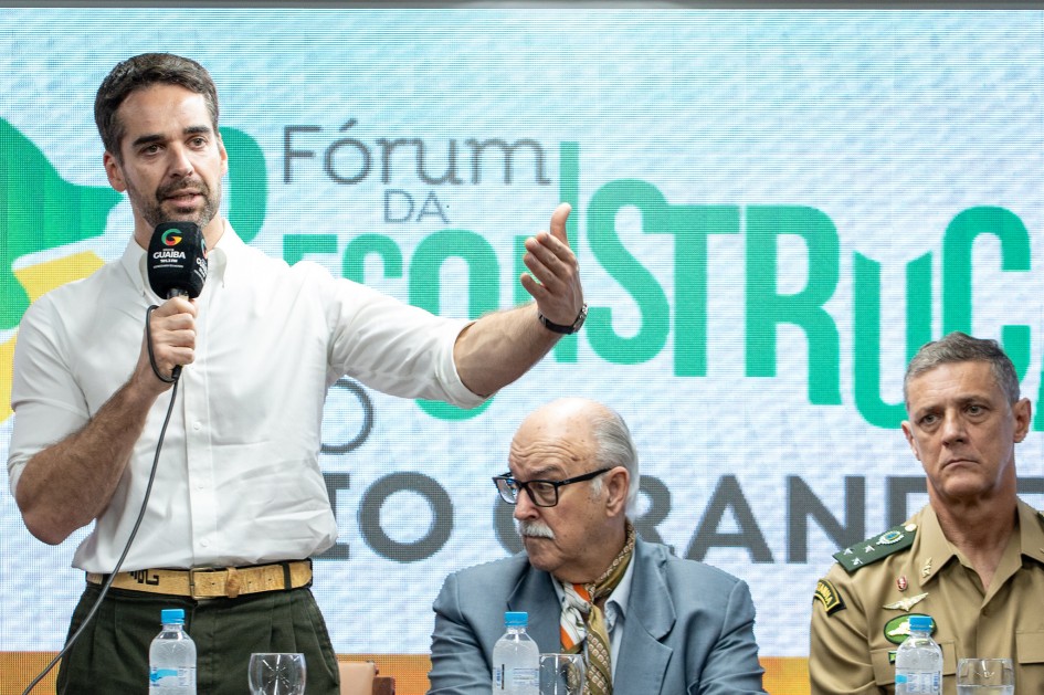 Em close, um homem de camisa branca fala ao microfone com o braço direito estendido, em gesto de ênfase, durante um evento oficial. À mesa à sua frente há garrafas de água e taças. Sentados ao lado, aparecem dois homens: um de terno azul-claro, óculos e lenço estampado no pescoço, que olha para o lado; e outro de farda bege com insígnias militares, que observa a fala. Ao fundo, o telão exibe a frase “Fórum da Reconstrução”.