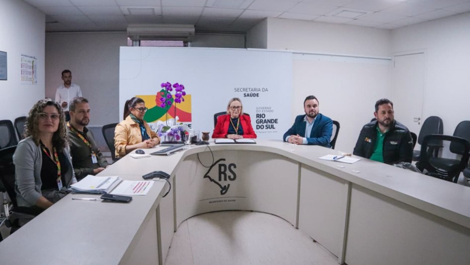 Foto em uma sala de reuniões, com três homens e três mulheres reunidos. A secretária da Saúde está posicionada ao centro.