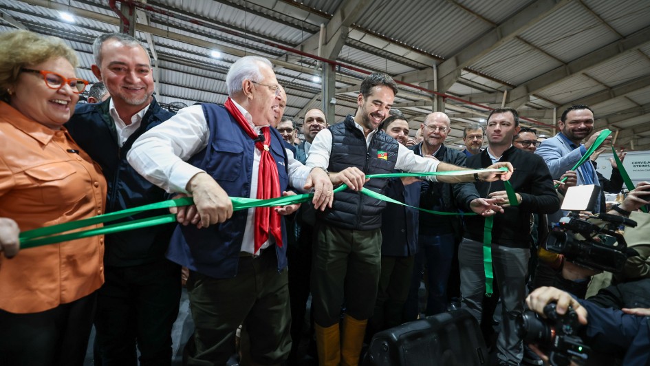 Em um pavilhão coberto, um grupo de autoridades e convidados participa de uma cerimônia de inauguração. No centro, um homem sorridente corta uma fita verde, segurada por pessoas ao seu redor, marcando a abertura oficial do espaço. Há fotógrafos registrando o momento e várias pessoas ao fundo acompanhando a solenidade. O clima é festivo e de celebração.