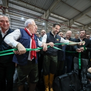 Em um pavilhão coberto, um grupo de autoridades e convidados participa de uma cerimônia de inauguração. No centro, um homem sorridente corta uma fita verde, segurada por pessoas ao seu redor, marcando a abertura oficial do espaço. Há fotógrafos registrando o momento e várias pessoas ao fundo acompanhando a solenidade. O clima é festivo e de celebração.