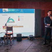 Um homem está de pé e fala ao microfone no canto direito da imagem. Ao fundo, há um telão com dados sobre os sorteados do Nota Fiscal Gaúcha de agosto. No palco, também há uma cadeira e instrumentos musicais. 