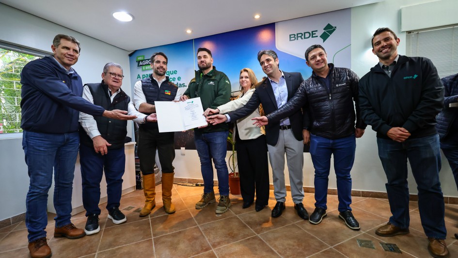 Foto colorida em ambiente interno mostra oito pessoas lado a lado posando para a câmera, com mais uma pessoa parcialmente visível à direita. No centro, dois homens seguram uma pasta aberta com um documento timbrado do BRDE; os demais estendem as mãos em direção ao papel e sorriem. Ao fundo, um painel com as marcas “BRDE” e o slogan “Meu Agro é BRDE”. O espaço tem piso de cerâmica, iluminação embutida no teto e janela com persianas à esquerda. Todas as pessoas vestem roupas de frio (jaquetas e coletes); um dos homens usa botas de couro de cano alto. Clima de celebração institucional.