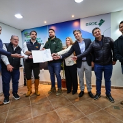 Foto colorida em ambiente interno mostra oito pessoas lado a lado posando para a câmera, com mais uma pessoa parcialmente visível à direita. No centro, dois homens seguram uma pasta aberta com um documento timbrado do BRDE; os demais estendem as mãos em direção ao papel e sorriem. Ao fundo, um painel com as marcas “BRDE” e o slogan “Meu Agro é BRDE”. O espaço tem piso de cerâmica, iluminação embutida no teto e janela com persianas à esquerda. Todas as pessoas vestem roupas de frio (jaquetas e coletes); um dos homens usa botas de couro de cano alto. Clima de celebração institucional.
