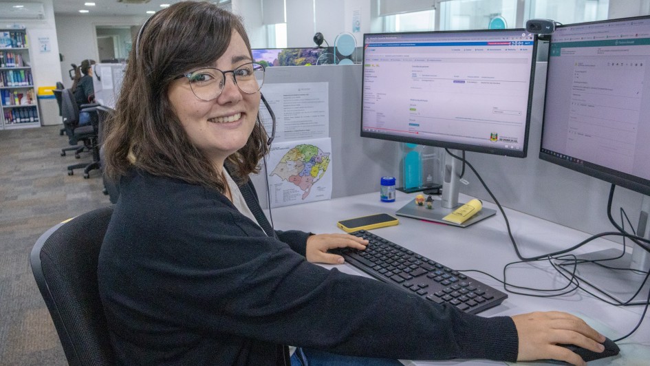 Uma mulher posa para a foto, sorrindo, em uma sala de trabalho. Ela está posicionada em frente a duas telas de computador.