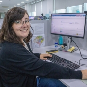 Uma mulher posa para a foto, sorrindo, em uma sala de trabalho. Ela está posicionada em frente a duas telas de computador. 