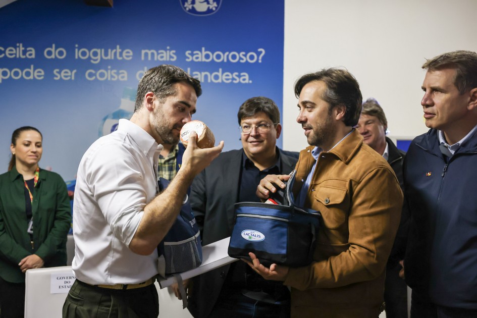 Governador Eduardo Leite aparece na foto cheirando um queijo pequeno redondo, que está na sua mão direita. Ele é observado por outros executivos. Um deles está à sua frente com uma bolsa azul aberta em suas mãos, com produtos lácteos.