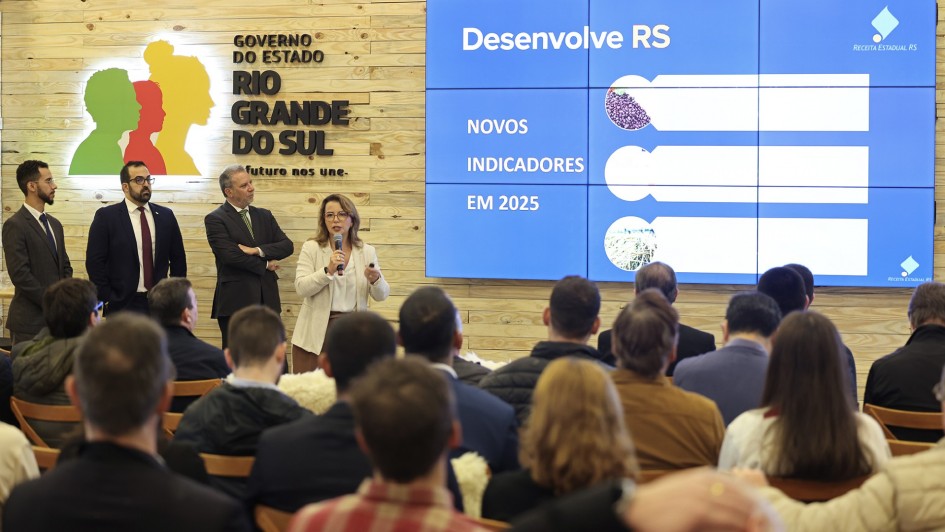 Foto mostra apresentação para uma plateia, que está de costas. Uma mulher, com casaco bege, segura o microfone e fala aos presentes. Três homens de terno estão ao seu lado. Há uma tela grande com imagem de fundo azul e o título Desenvolve RS Novos Indicadores em 2025. Há também um logo do governo estadual.