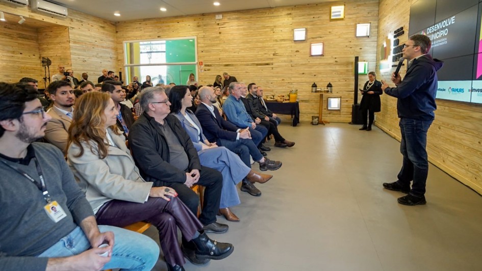 Foto mostra palestrante numa sala com paredes em madeira clara, em pé, com um microfone na mão. Ele está diante de uma plateia que o acompanha atentamente.