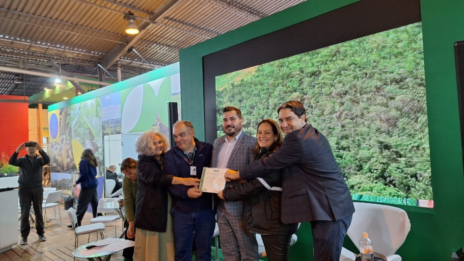 Foto mostra um grupo de cinco pessoas que seguram um documento do certificado na mão. São três homens de terno e duas mulheres. Eles estão num estande da feira, na frente de um telão que exibe uma imagem de uma floresta.