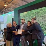 Foto mostra um grupo de cinco pessoas que seguram um documento do certificado na mão. São três homens de terno e duas mulheres. Eles estão num estande da feira, na frente de um telão que exibe uma imagem de uma floresta. 