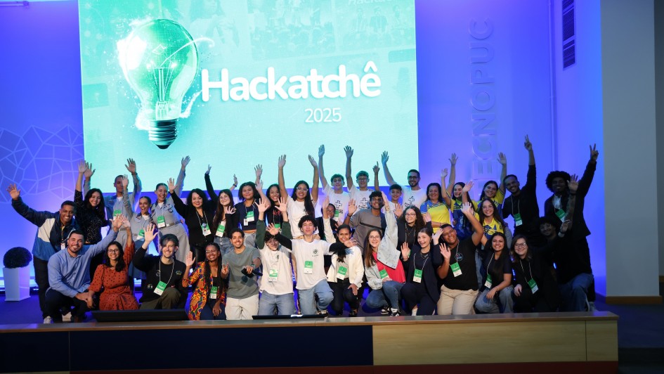 Um grupo de pessoas está reunido em cima de um palco com as mãos para cima, posando para a foto. Ao fundo, no telão, lê-se "HackaTchê 2025". 