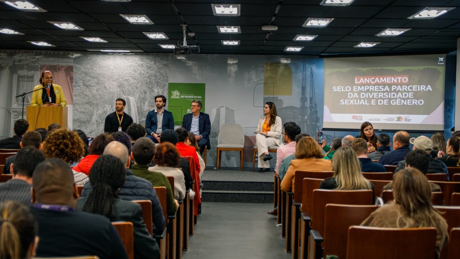 Fotografia de um auditório com cadeiras ocupadas por diversas pessoas, todas voltadas para o palco. No palco, há uma mesa com quatro pessoas sentadas — duas mulheres e dois homens — e, ao lado, uma mulher em pé, falando ao microfone em um púlpito de madeira. No fundo, um telão exibe a mensagem: “Lançamento – Selo Empresa Parceira da Diversidade Sexual e de Gênero”. Atrás do palco, há um painel com imagens e o logotipo do Governo do Estado do Rio Grande do Sul. O ambiente é iluminado e transmite o clima de um evento formal de lançamento.