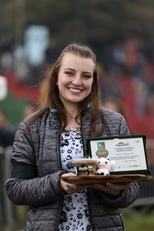 Uma jovem sorri para a câmera, segurando uma bandeja de madeira. Na bandeja, há um troféu verde com inscrições e um pequeno objeto decorativo de uma vaca. A jovem usa uma jaqueta cinza com detalhes pretos e uma blusa estampada. Ao fundo, as pessoas assistem a um evento. A imagem celebra uma vencedora da Expointer 2025.