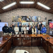 A foto mostra um grupo de aproximadamente 15 pessoas em pé atrás de uma grande mesa em formato de U. Todos parecem estar participando de um evento de assinatura de parceria. No lado esquerdo da imagem, há um telão com o logo da UNISINOS. Na parede ao fundo, há quadros com imagens rústicas e um banner colorido da PRF com o título "CONHEÇA OS PRINCIPAIS PROJETOS". A sala tem paredes de pedra e madeira no teto, com iluminação artificial forte. As pessoas vestem uma mistura de roupas casuais e uniformes, incluindo alguns da Polícia Rodoviária Federal (PRF). Uma mulher no centro do grupo está segurando um documento, possivelmente um acordo ou termo de parceria.