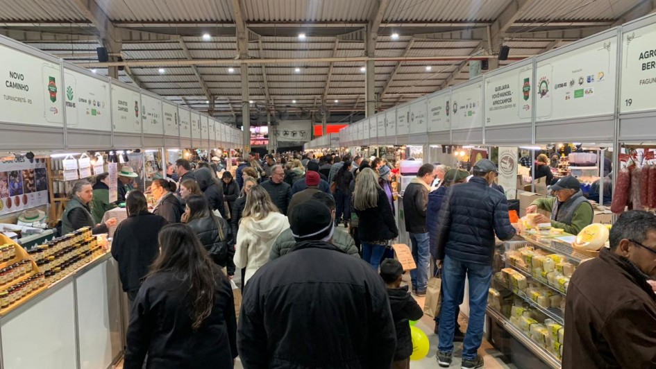 Descrição para fins de acessibilidade: Fotografia mostra o interior do Pavilhão da Agricultura Familiar da Expointer, repleto de visitantes. Pessoas circulam pelos corredores e formam filas em frente aos estandes para conhecer e comprar produtos como queijos, salames e geleias. Os estandes estão identificados com placas brancas, e o ambiente apresenta grande movimento, evidenciando a forte presença do público no local.