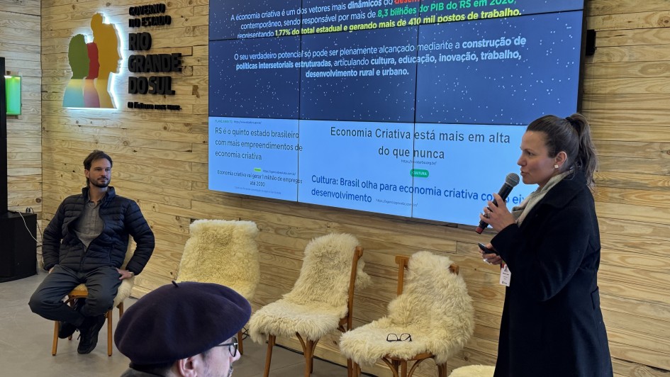Descrição para fins de acessibilidade: Em uma sala com paredes de madeira clara, uma mulher fala ao microfone diante de um telão que exibe dados sobre economia criativa no Rio Grande do Sul. Ao fundo, há três cadeiras com mantas de lã branca. Um homem sentado observa a apresentação, enquanto outra pessoa aparece parcialmente em primeiro plano, usando boina e óculos. Na parede, à esquerda, está o logotipo colorido do Governo do Estado do Rio Grande do Sul.