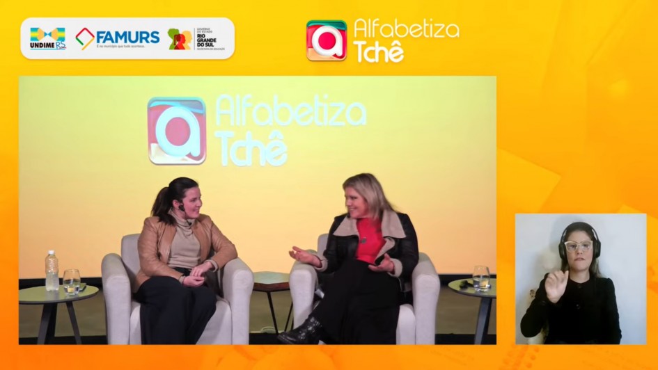 Imagem de um evento do programa Alfabetiza Tchê. Duas mulheres estão sentadas em poltronas no palco, conversando entre si. À esquerda, uma mesa lateral com garrafa de água e copo. No canto inferior direito da tela, aparece uma janela com uma intérprete de Libras usando fones de ouvido e óculos. O fundo do palco é amarelo, com os logotipos da Undime-RS, Famurs e do Governo do Estado do Rio Grande do Sul – Secretaria da Educação, além da identidade visual do programa Alfabetiza Tchê.