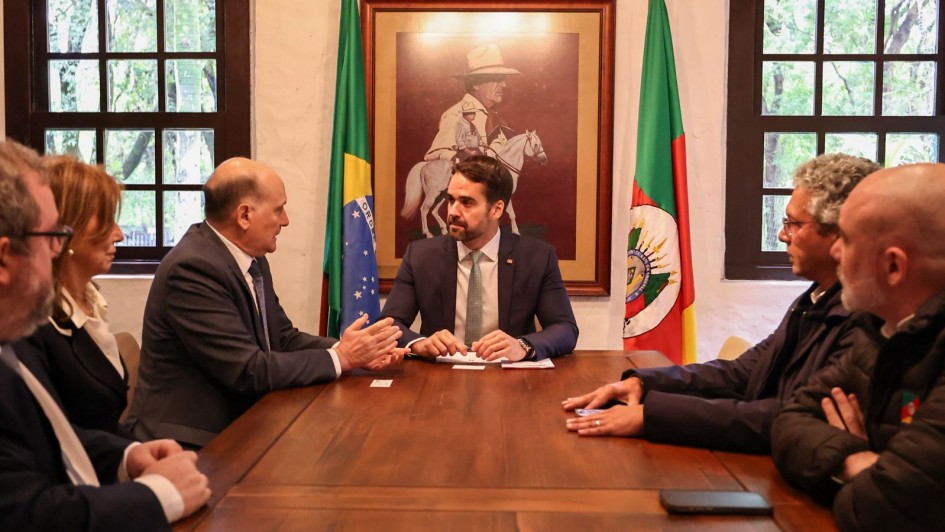 Governador está conversando com o embaixador, eles estão sentados em volta de uma mesa de madeira. Outras pessoas estão sentadas acompanhando a conversa. Ao fundo, a bandeira do Brasil e do Rio Grande do Sul.