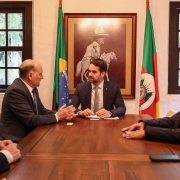 Governador está conversando com o embaixador, eles estão sentados em volta de uma mesa de madeira. Outras pessoas estão sentadas acompanhando a conversa. Ao fundo, a bandeira do Brasil e do Rio Grande do Sul.