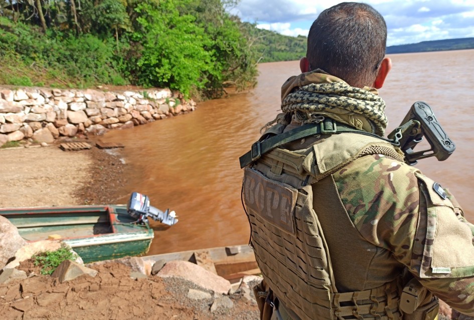 Policial com roupa camuflada e colete tático está de pé à beira de um rio barrento, olhando para a água. Ao lado, há um pequeno barco verde e pedras grandes na margem. O ambiente é cercado por árvores e vegetação.