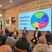 Foto mostra cinco painelistas sentados diante da plateia. O homem do centro está falando ao microfone. Atrás dele há um telão onde está escrito Diálogos Energia e Futuro e um gráfico em circulo, não legível na foto. Ao lado do telão, a logomarca do RS. 