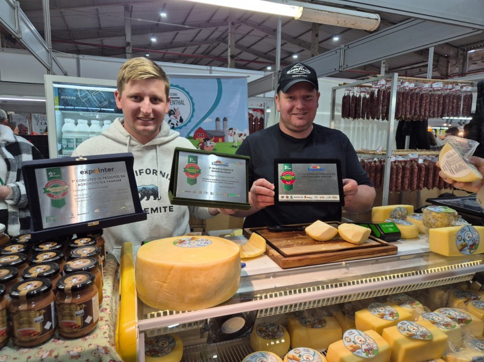 Foto mostra dois produtores em sua banca. Ele seguram três placas recebidas em premiações de seus produtos. Na banca estão em destaque um grande queijo amarelo inteiro e outros menores cortados, além de geleias e outros produtos colonais. Ao fundo, um suporte com dezenas de salames pendurados.