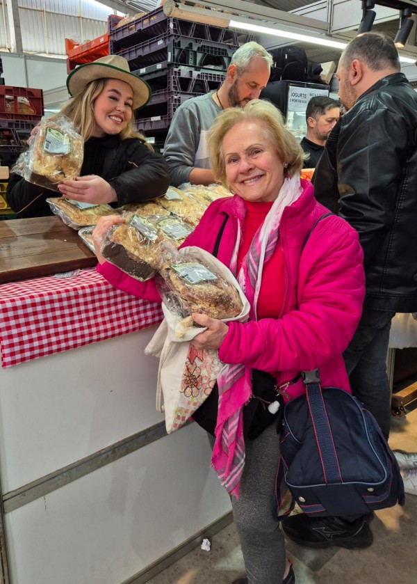 Foto mostra Nilva Fransceschini na banca Nona Diva, segurando uma cuca um bolo. Atrás dela há uma banca onde outra mulher segura outra cuca. Há outro desses bolos sobre a bancada. Atrás há outros homens sendo atendidos em outra banca.