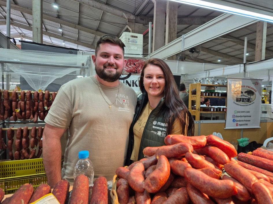 Foto mostra casal de produtores, em sua banca de salames. Há salames sobre a banca, em primeiro plano. E salames, ao fundo, pendurados em um varal.