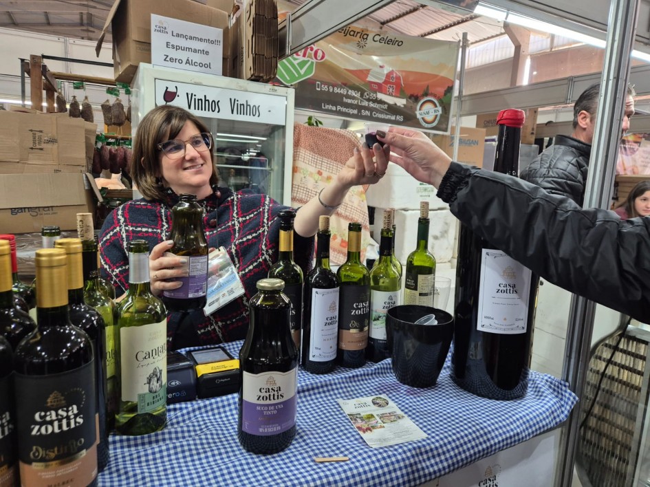 Foto mostra produtora em sua banca de vinhos e sucos de uva. Ela segura uma garrafa e serve um cliente, que só aparece o braço.