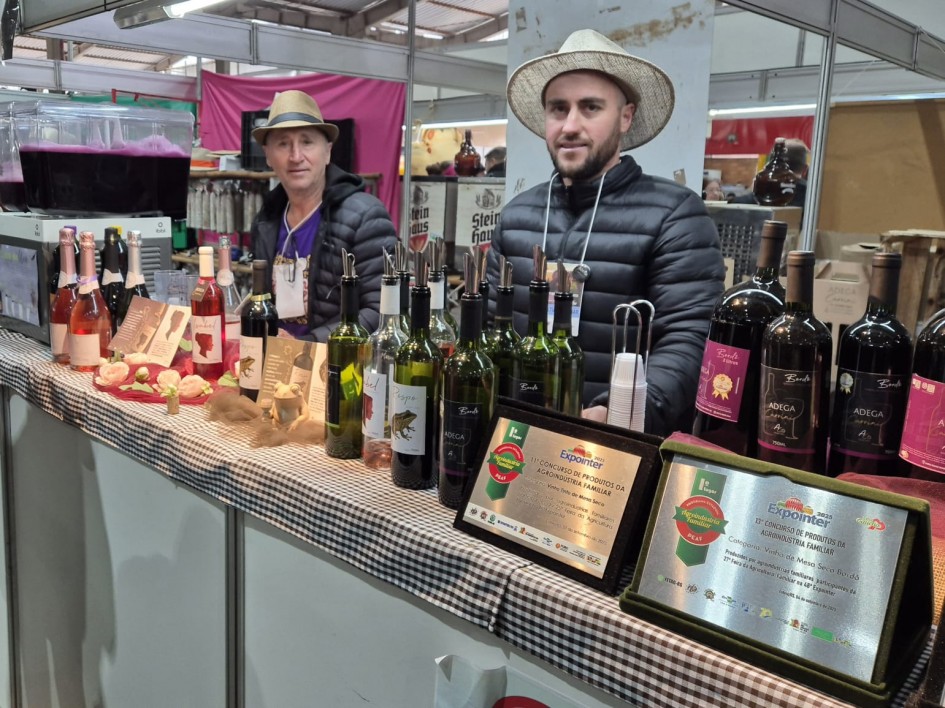 Dois produtores estão atrás da bancada de sua banca, sobre a qual estão dezenas de garrafas de vinhos, de diferentes variedades. Sobre a mesa, duas placas de premiações recebidas pelos produtos.
