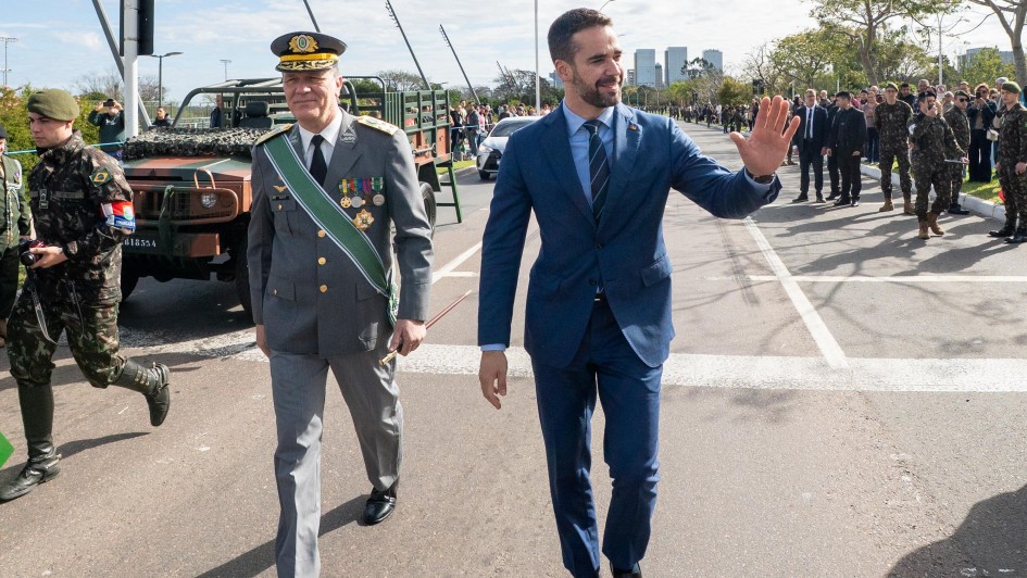 Nesta foto, o governador Eduardo Leite, em um terno azul, acena para o público, demonstrando receptividade e engajamento durante o Desfile de 7 de Setembro em Porto Alegre. Ao seu lado, um oficial militar em uniforme de gala acompanha o momento. A presença de militares e equipamentos ao fundo reforça o contexto do evento cívico-militar.