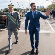 Nesta foto, o governador Eduardo Leite, em um terno azul, acena para o público, demonstrando receptividade e engajamento durante o Desfile de 7 de Setembro em Porto Alegre. Ao seu lado, um oficial militar em uniforme de gala acompanha o momento. A presença de militares e equipamentos ao fundo reforça o contexto do evento cívico-militar.