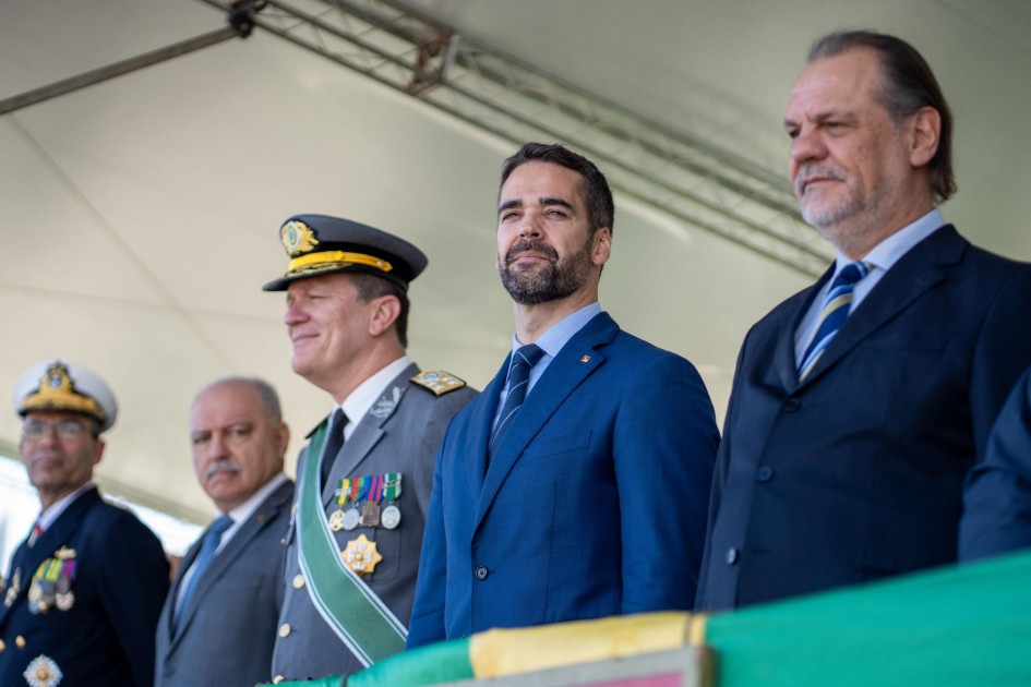 A imagem exibe uma fileira de autoridades em um palanque, destacando a presença do governador Eduardo Leite. Ele, vestindo um terno azul, está posicionado no centro, com um semblante sério e atento. Ao seu lado, militares em trajes de gala e outros representantes civis compõem a cena, transmitindo a importância do evento cívico-militar. O fundo, com um céu claro e uma estrutura de cobertura, em um evento oficial em celebração ao Dia da Independência.