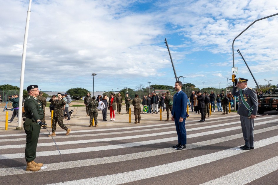 A imagem foca em um momento de formalidade durante o Desfile de 7 de Setembro em Porto Alegre, com o governador Eduardo Leite em trajes civis e um oficial militar em uniforme de gala. Eles se encontram em uma faixa de pedestres, com outras pessoas e veículos militares ao fundo. A cena sugere um momento de revista das tropas antes do desfile cívico-militar.