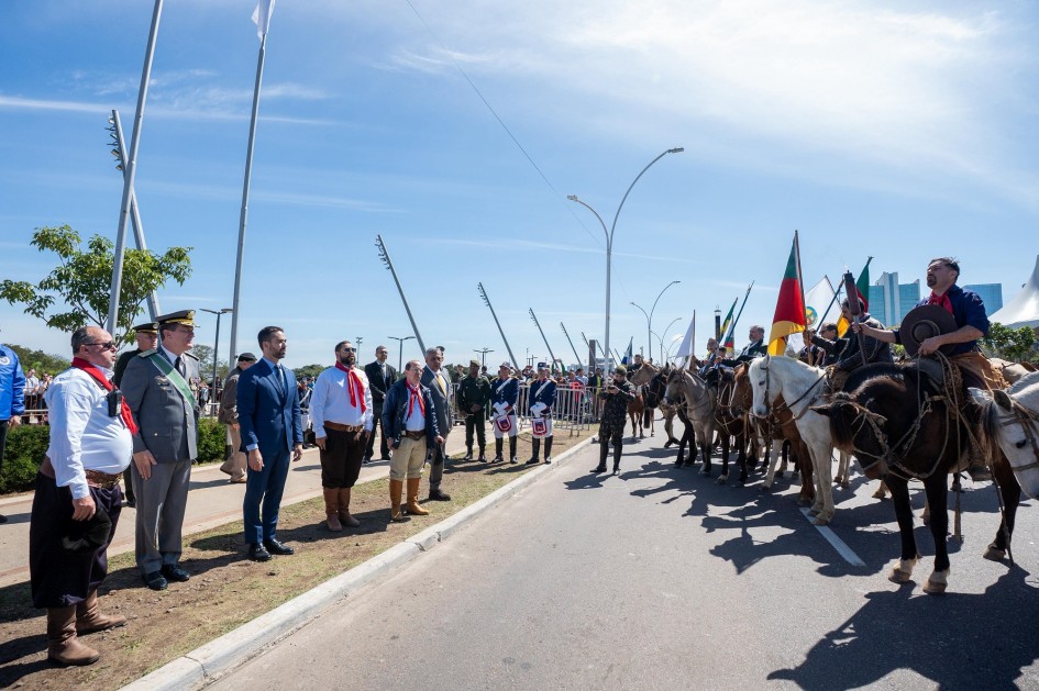 A fotografia captura um momento do Desfile de 7 de Setembro em Porto Alegre, onde figuras importantes, incluindo o governador Eduardo Leite, observam a passagem de participantes a cavalo. Os cavaleiros, trajando vestimentas que remetem à tradição gaúcha e empunhando bandeiras, trazem um elemento cultural distinto à celebração. A linha do horizonte com prédios ao longe e o céu azul indicam o cenário urbano e o bom tempo para o evento.