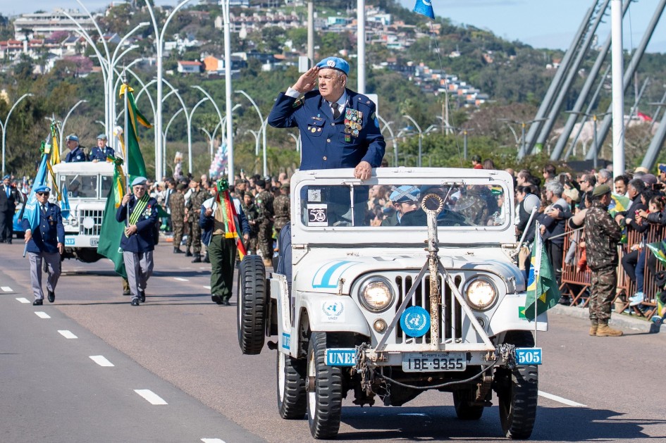 A cena mostra um militar em um jipe, possivelmente em um momento de saudação ou reconhecimento durante o Desfile de 7 de Setembro em Porto Alegre. O militar, com um boné azul da ONU e distintivos, está em destaque, em frente a outros veículos e participantes do desfile. A multidão à beira da estrada e os prédios ao fundo indicam a presença de espectadores e o ambiente da cidade.