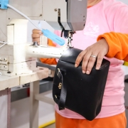 Pessoa costura uma bolsa de couro preta em máquina industrial. Ela usa roupa colorida e guia o material com precisão. A bolsa tem fivela dourada e está sendo montada com cuidado.