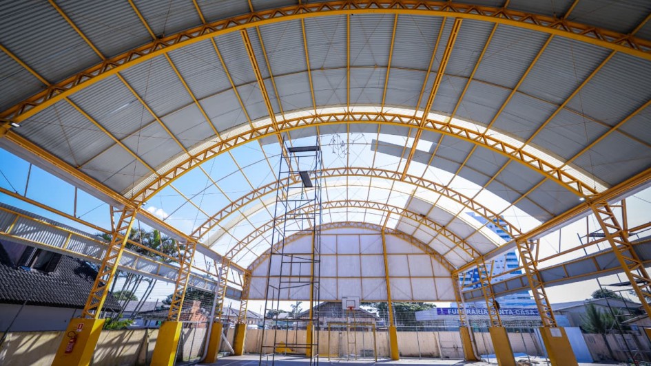 A imagem mostra o interior de uma quadra poliesportiva coberta, com uma estrutura metálica de teto em formato de arco. A cobertura translúcida permite a entrada de luz natural, enquanto as telhas metálicas garantem proteção. Andaimes e materiais de construção espalhados indicam que o local está passando por reformas ou manutenção. A presença de uma cesta de basquete e as cores da estrutura remetem a um ambiente esportivo e escolar.