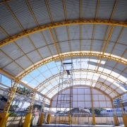 A imagem mostra o interior de uma quadra poliesportiva coberta, com uma estrutura metálica de teto em formato de arco. A cobertura translúcida permite a entrada de luz natural, enquanto as telhas metálicas garantem proteção. Andaimes e materiais de construção espalhados indicam que o local está passando por reformas ou manutenção. A presença de uma cesta de basquete e as cores da estrutura remetem a um ambiente esportivo e escolar.