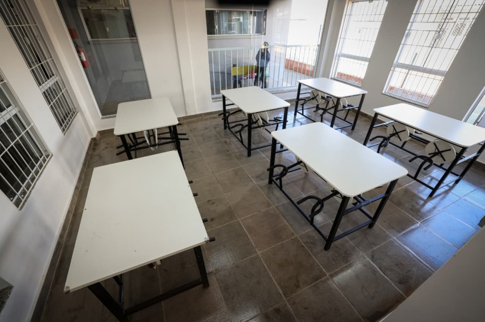 Esta imagem mostra um refeitório com várias mesas brancas dispostas em um padrão organizado. As mesas possuem estruturas metálicas escuras e tampos brancos. As paredes são claras, e há janelas com grades metálicas, além de portas.