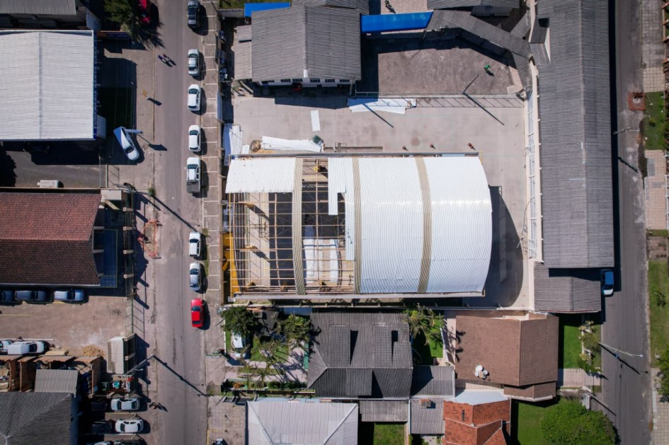 Esta imagem aérea oferece uma visão detalhada de uma área urbana, mostrando a disposição de casas com telhados de barro e cinza, ruas com carros estacionados e uma quadra poliesportiva em construção. A cobertura da quadra está em fase final, com a estrutura metálica aparente e telhas sendo instaladas. Ao redor, outras construções residenciais e comerciais completam a paisagem.