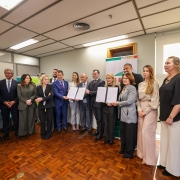 Foto mostra várias pessoas, vestidas formalmente, posam para a foto segurando documentos. Destaca-se uma mulher com um vestido preto e outra com um terno cinza, ambas segurando papéis assinados. 