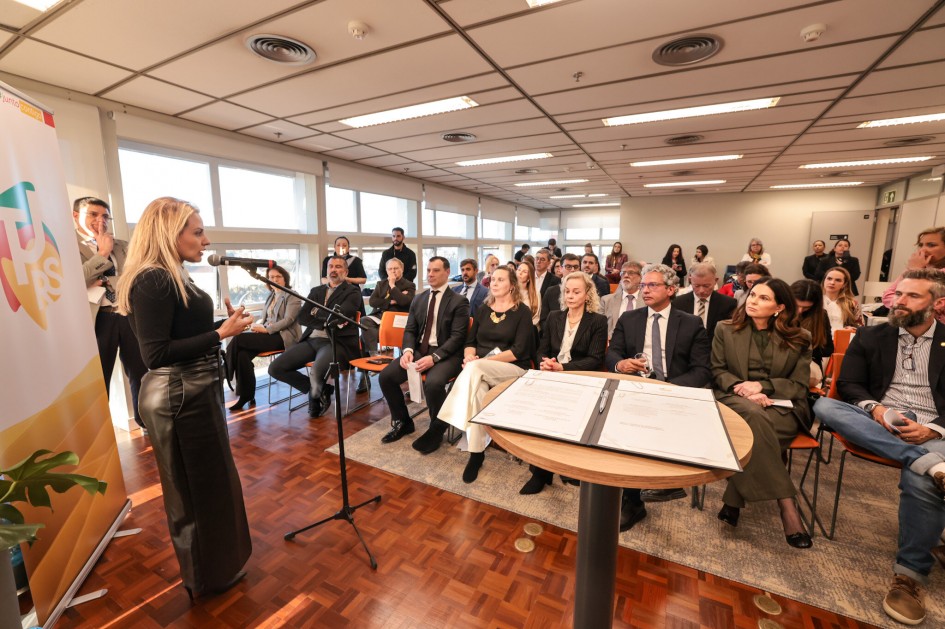 Nesta foto, a secretária de Planejamento, Governança e Gestão, Danielle Calazans, discursa em pé, em frente a um púlpito, com um microfone. O ambiente, com janelas amplas e um banner ao fundo. Uma plateia está sentada à sua frente e observa atentamente.