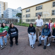 O governador Eduardo Leite está em um pátio escolar, conversando com um grupo de estudantes que estão sentados em cadeiras, em um círculo. Ele gesticula enquanto fala, indicando que está explicando algo ou ouvindo atentamente. Ao fundo, o prédio do colégio e outros edifícios residenciais compõem a paisagem.