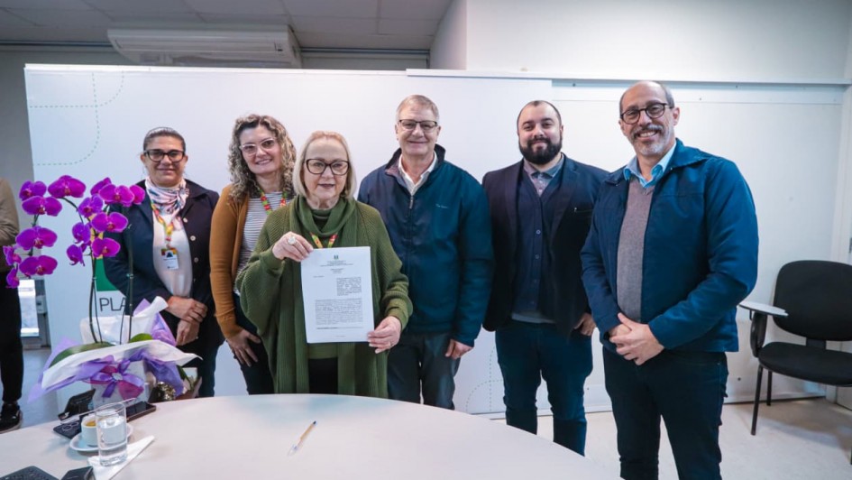 Seis pessoas est&atilde;o em p&eacute;, lado a lado, sorrindo para a foto em um ambiente interno iluminado, provavelmente uma sala de reuni&otilde;es. No centro, uma mulher mais velha, de &oacute;culos e vestindo um casaco verde com cachecol, segura um documento assinado &mdash; sugerindo um momento de formaliza&ccedil;&atilde;o, como a assinatura de um conv&ecirc;nio. &Agrave; esquerda, h&aacute; um vaso com flores roxas sobre a mesa redonda clara, onde tamb&eacute;m est&atilde;o um copo com &aacute;gua, um telefone e uma caneta. As pessoas ao redor vestem roupas formais e casuais, e o ambiente transmite um clima profissional e cordial.