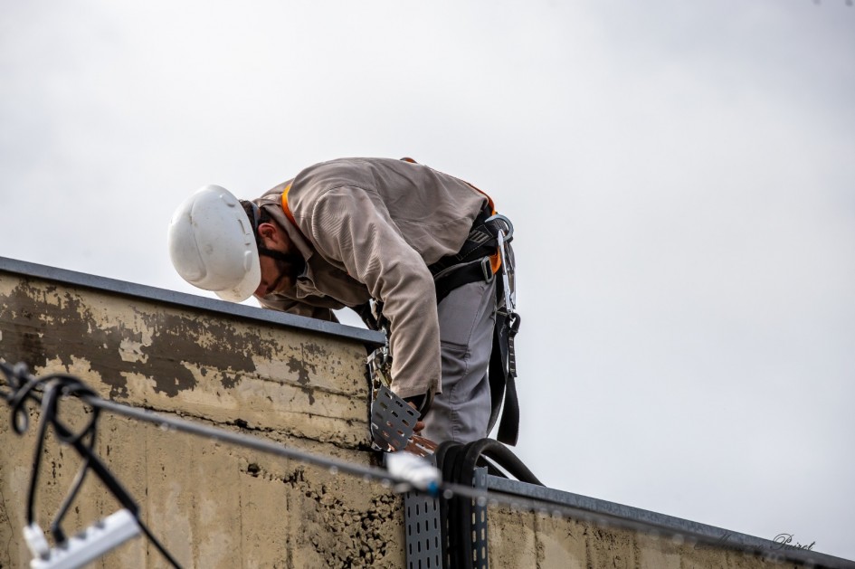 A imagem mostra um trabalhador da construção civil em cima de uma edificação. Ele está curvado sobre a borda de um telhado, aparentemente realizando algum tipo de instalação ou manutenção. O trabalhador usa equipamentos de segurança, incluindo capacete branco, cinto de segurança com talabarte e uniforme de proteção cinza. Na borda do telhado, há sinais de desgaste e deterioração do concreto. Também é possível ver cabos e suportes metálicos ligados à estrutura. O céu está nublado ao fundo, indicando um dia sem sol.