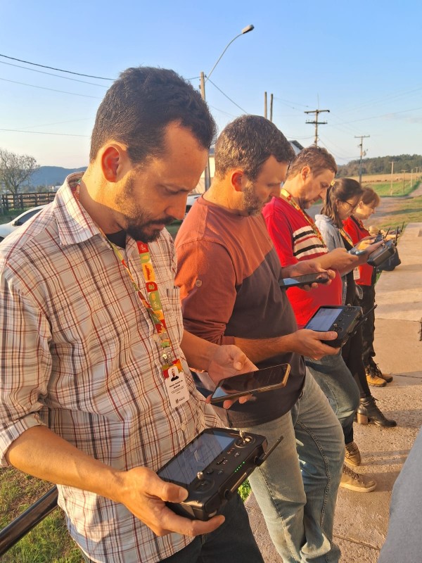 A imagem mostra um grupo de cinco pessoas em pé ao ar livre, alinhadas lado a lado, cada uma segurando controles remotos de drones. Quatro delas também utilizam smartphones, apoiados nas mãos junto ao equipamento de controle. Todas parecem concentradas, olhando para os dispositivos. O cenário é iluminado pela luz do entardecer, com postes, fiação elétrica e campos ao fundo. 